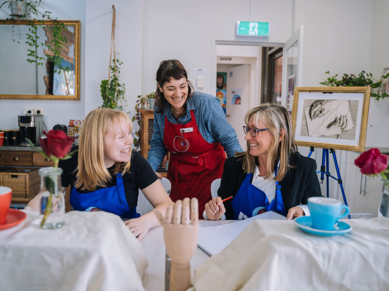 A teacher smiling at two women at an art class.